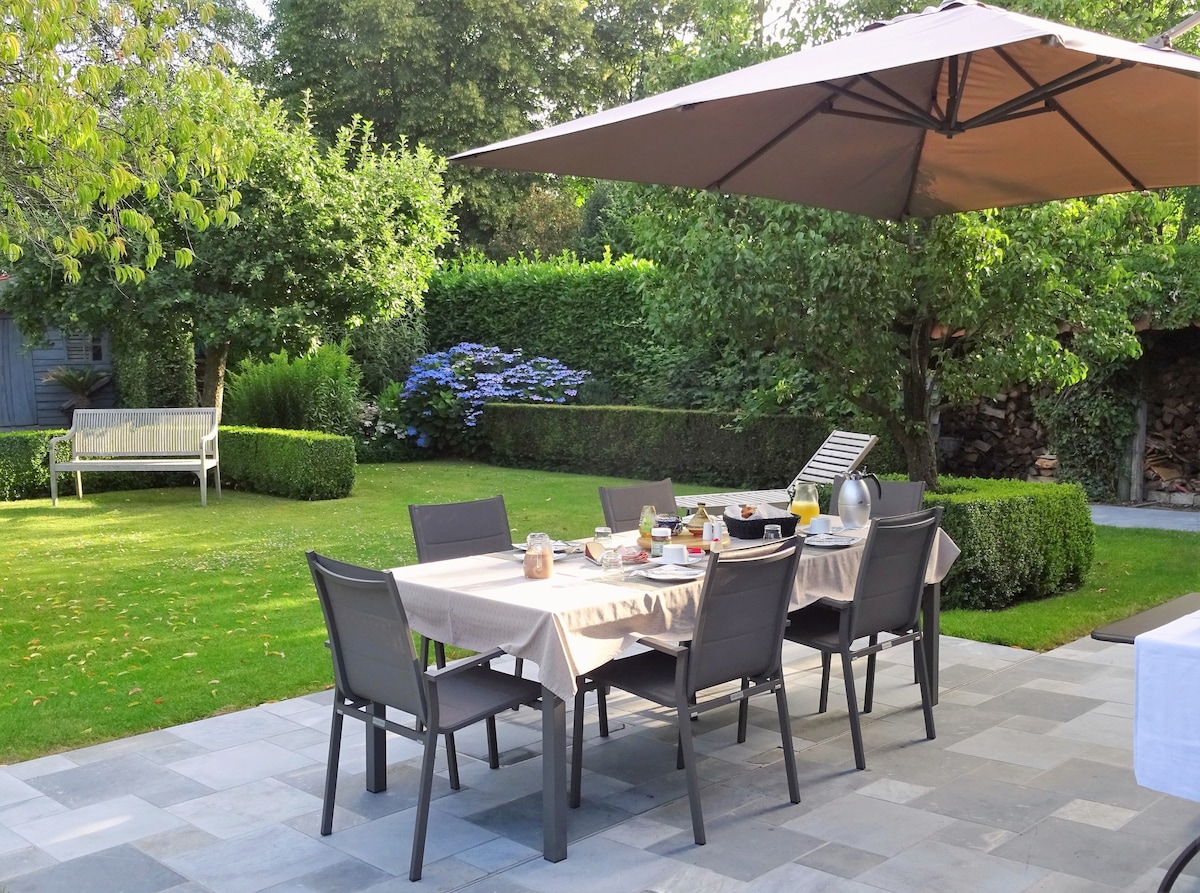 A spacious outdoor dining area is set with a large table and comfortable chairs beneath a shaded umbrella. The setting is surrounded by lush greenery, featuring flowering plants and neatly trimmed hedges. A sun lounger is positioned nearby on the well-maintained lawn.