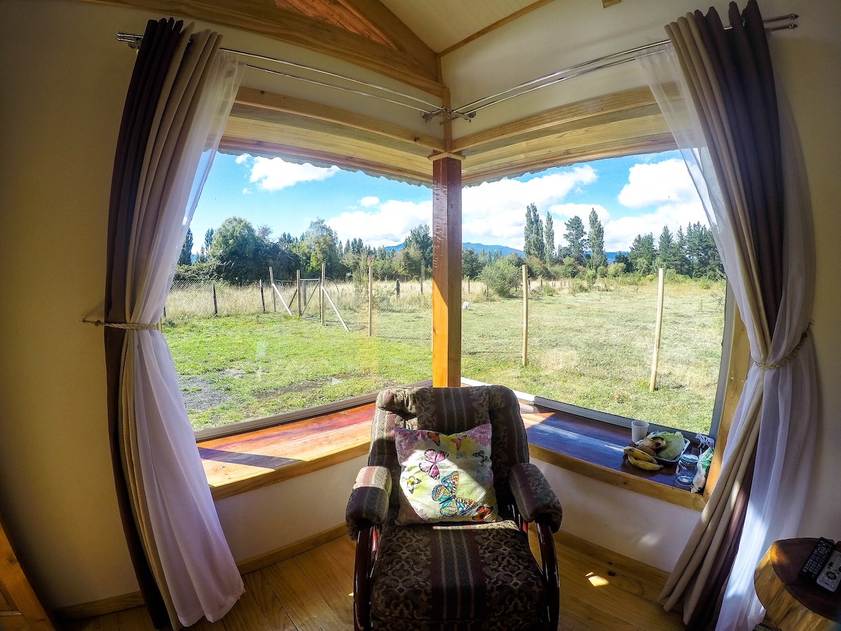 A cozy seating area is highlighted by a bow window, providing a panoramic view of the surrounding grassy landscape and distant mountains. Light drapes frame the window, enhancing the natural brightness of the space.