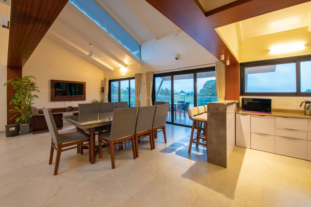 A spacious dining area features a large wooden table surrounded by gray upholstered chairs. Sunlight streams through expansive glass doors, connecting the indoor space to the outdoor balcony, while a modern kitchen setup is visible in the background.