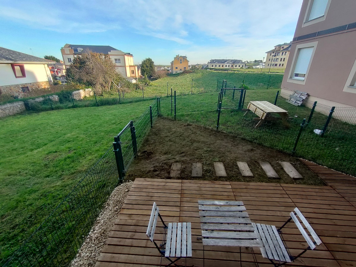 An outdoor space is shown, featuring a wooden deck and simple seating arrangements. A fenced-in grassy area extends beyond, providing an unobstructed view of the surrounding landscape. Nearby structures are visible in the distance, contributing to the peaceful setting.