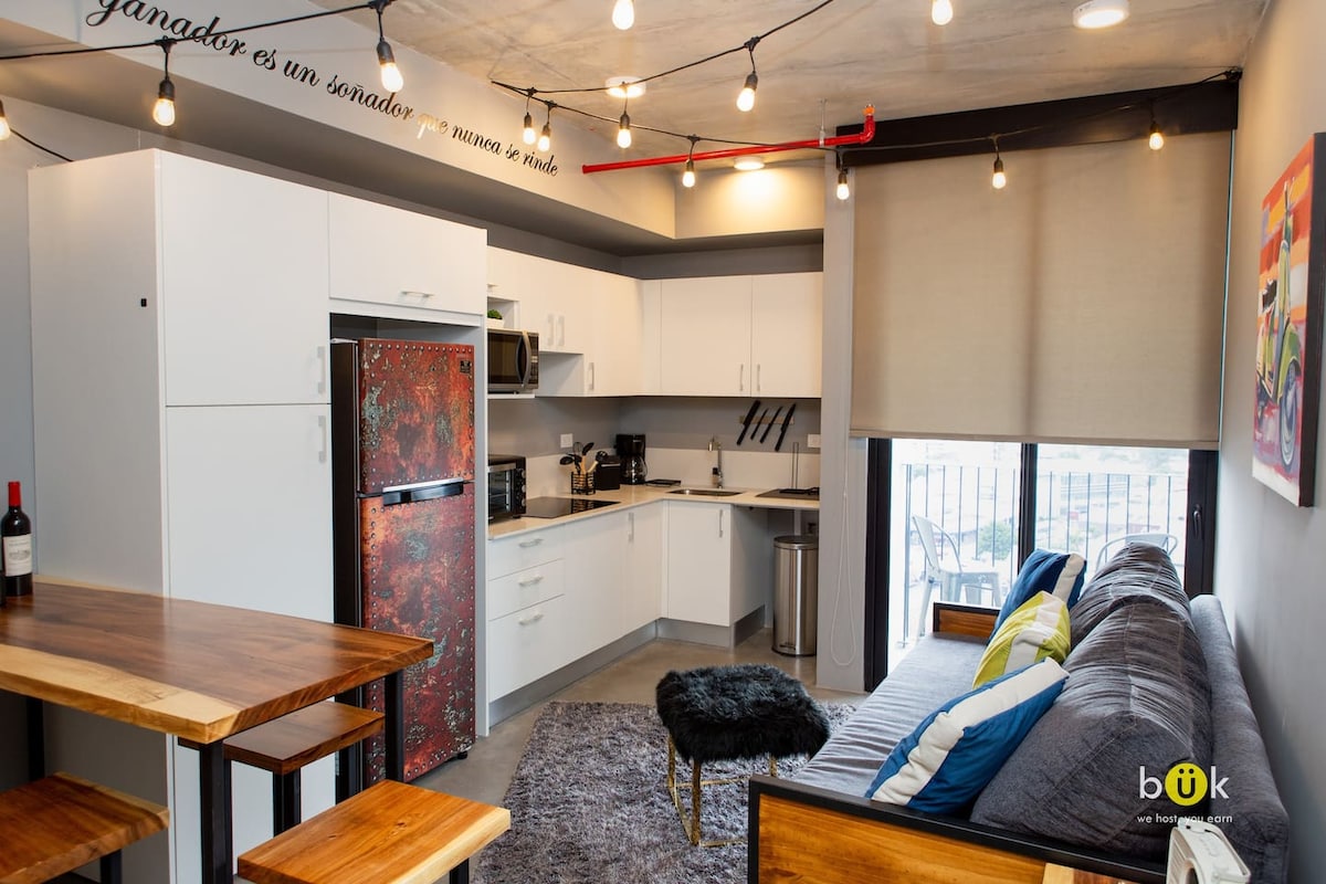 A modern kitchen is paired with an inviting living area, featuring a comfortable sofa and a wooden breakfast bar. The space is well-lit by decorative string lights above and natural light filtering through large windows. Appliances, including a refrigerator and microwave, are neatly arranged.