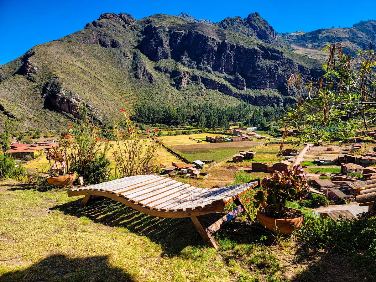 A wooden lounge chair is positioned on a grassy terrace, offering a view of rolling hills and terraced fields below. Vibrant flowers are placed in pots alongside the chair, adding a touch of color to the serene outdoor space.