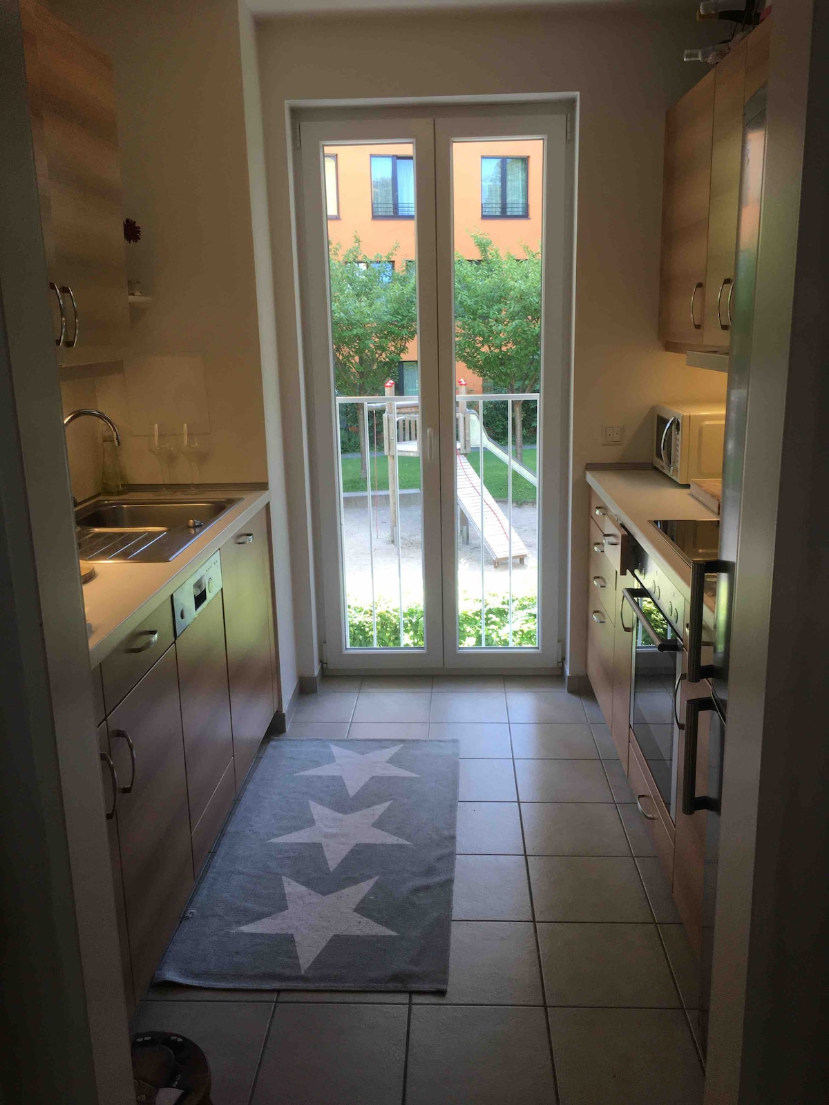 A compact kitchen space features wooden cabinetry and modern appliances, including an oven and microwave. Natural light enters through double glass doors that open to a small balcony overlooking greenery. A light-colored rug with star patterns adds a subtle touch to the tiled floor.