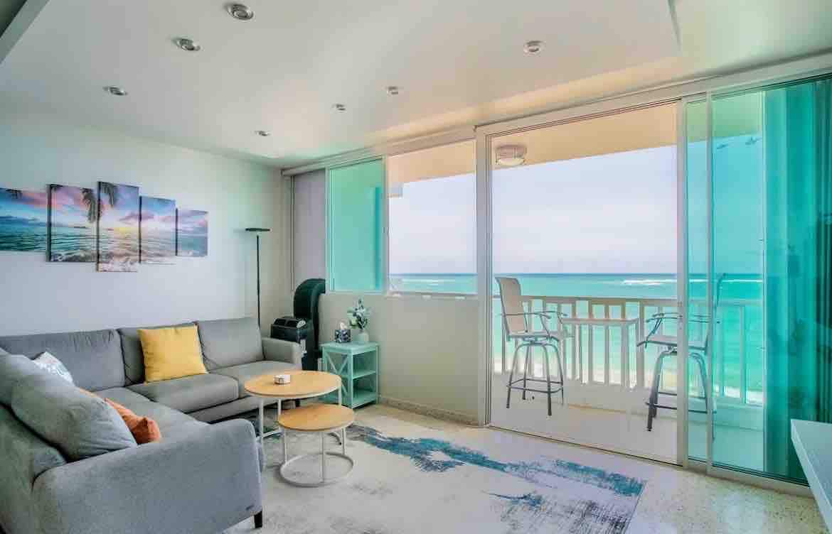 A modern living area features a plush gray sofa with yellow and white accents, facing large glass doors that open to the ocean view. A light blue table sits between the sofa and a small dining area with tall chairs, framing the coastal landscape beyond.