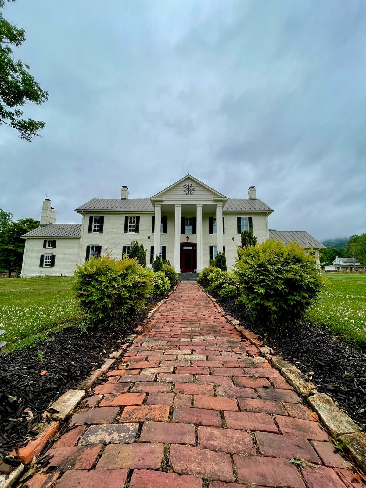 A charming brick pathway leads to the grand facade of the Manor House, featuring white columns and dark shutters. Lush green bushes frame the entrance, while the expansive lawn offers a serene setting under a cloudy sky.