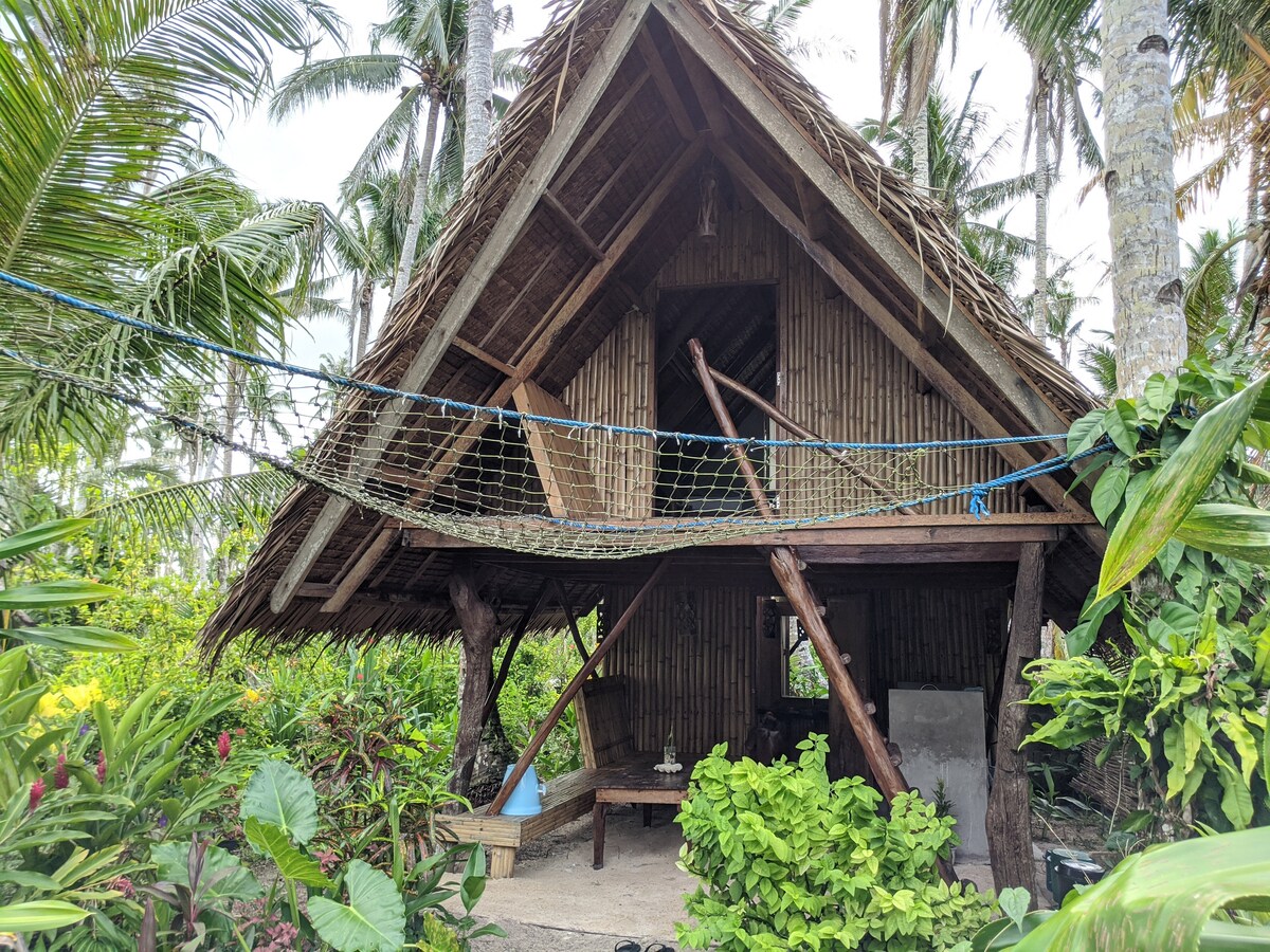 A traditional bamboo treehouse is surrounded by lush greenery, featuring a thatched roof and wooden beams. A large hammock net is strung between the posts, and tropical plants add vibrant colors to the scene. The natural setting conveys a sense of tranquility and connection to nature.