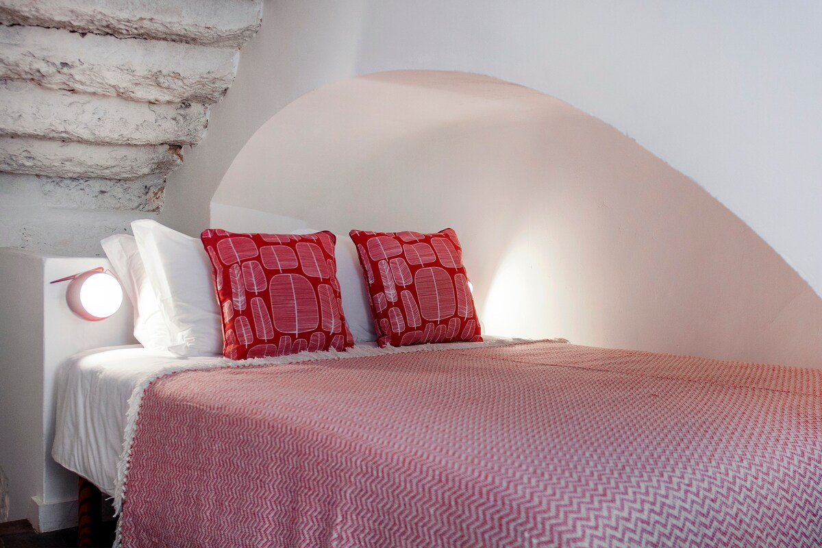 Შეაბიჯეთ ამ მყუდრო ოთახში, სადაც საწოლი ნაწილობრივ მოთავსებულია მომხიბვლელ არკადში, რბილი ტექსტურებითა და თბილი წითელი აქცენტებით, რაც კომფორტის იდეალურ თავშესაფარს ქმნის.