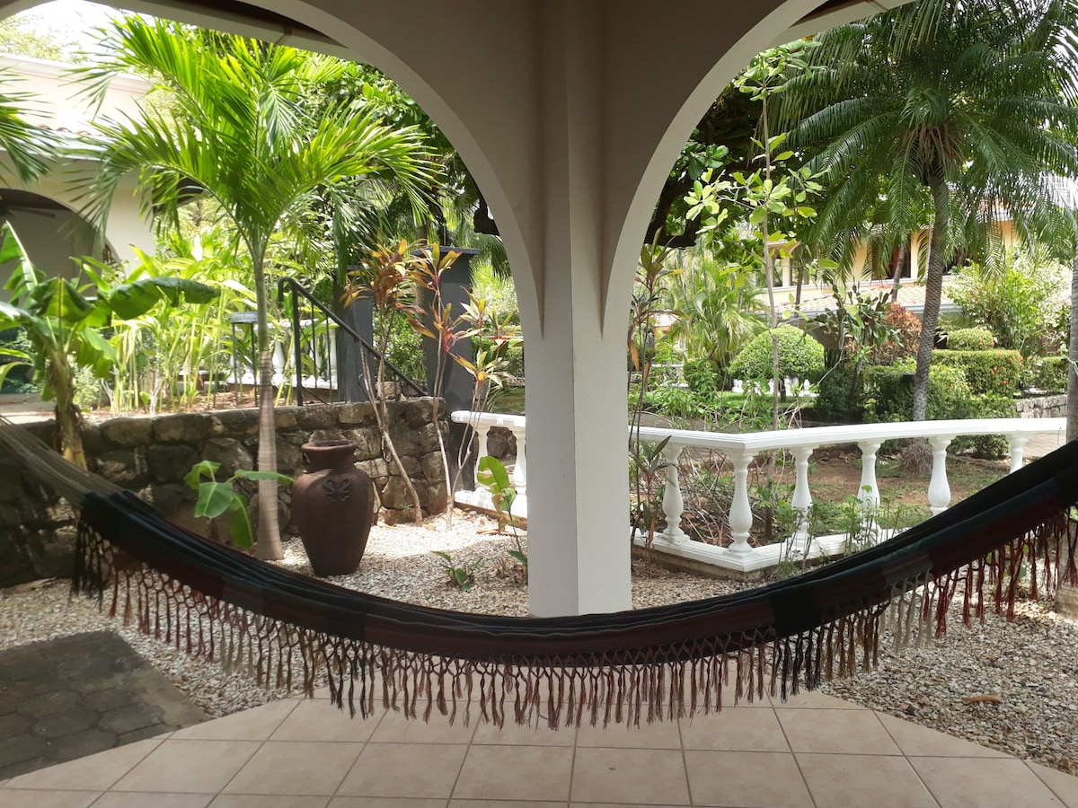 A hammock is suspended between two columns, inviting relaxation in a shaded area. Tropical plants surround the space, with a stone pathway leading to a staircase. Decorative terracotta pots are positioned among the greenery, adding to the tranquil ambiance.