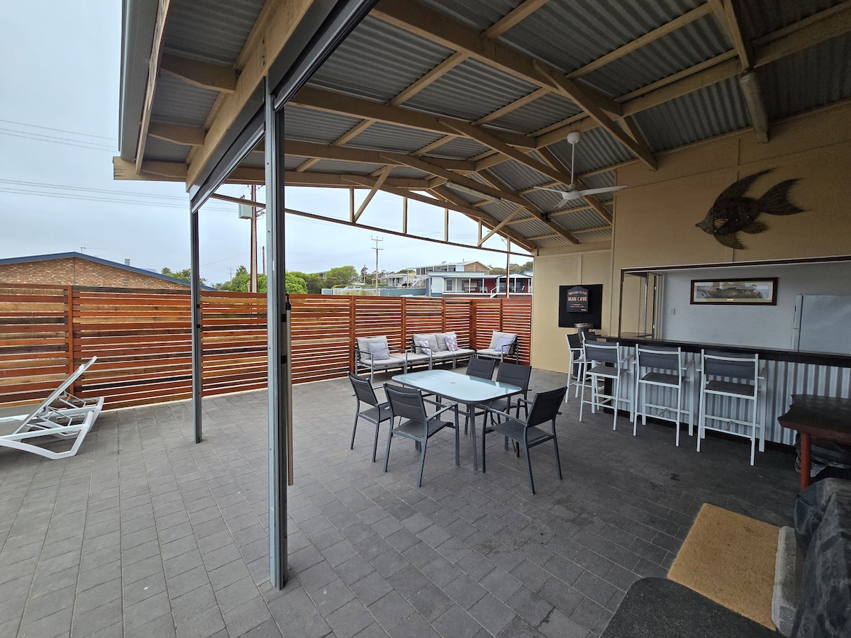 The large undercover patio features a spacious dining table surrounded by several chairs. An outdoor bar with seating is visible, along with a lounge area and a wall-mounted television, providing an inviting space for gatherings. The area is enclosed with timber fencing.