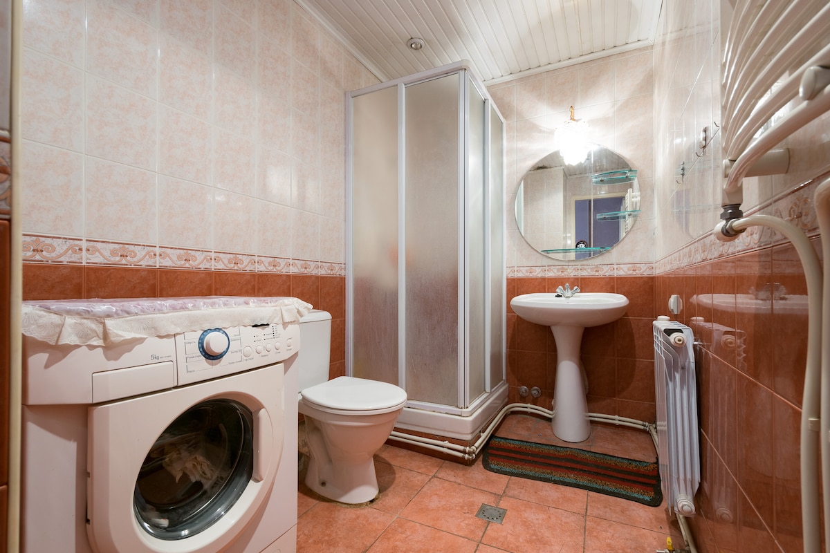 A full bathroom is displayed, featuring a shower with frosted glass doors, a round mirror above a pedestal sink, and a modern washing machine. The space is finished with tiled walls and a patterned rug on the floor, providing a functional and clean environment.
