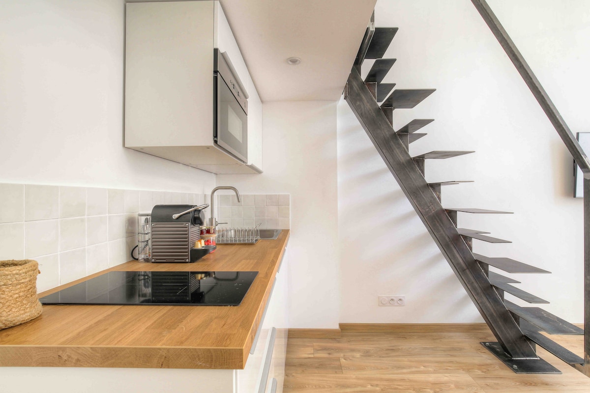 A modern kitchenette is featured with a sleek wooden countertop and built-in appliances, including a microwave and an induction cooktop. A stylish, open staircase with metal steps leads to the upper level, while light wood flooring adds a warm touch to the space.
