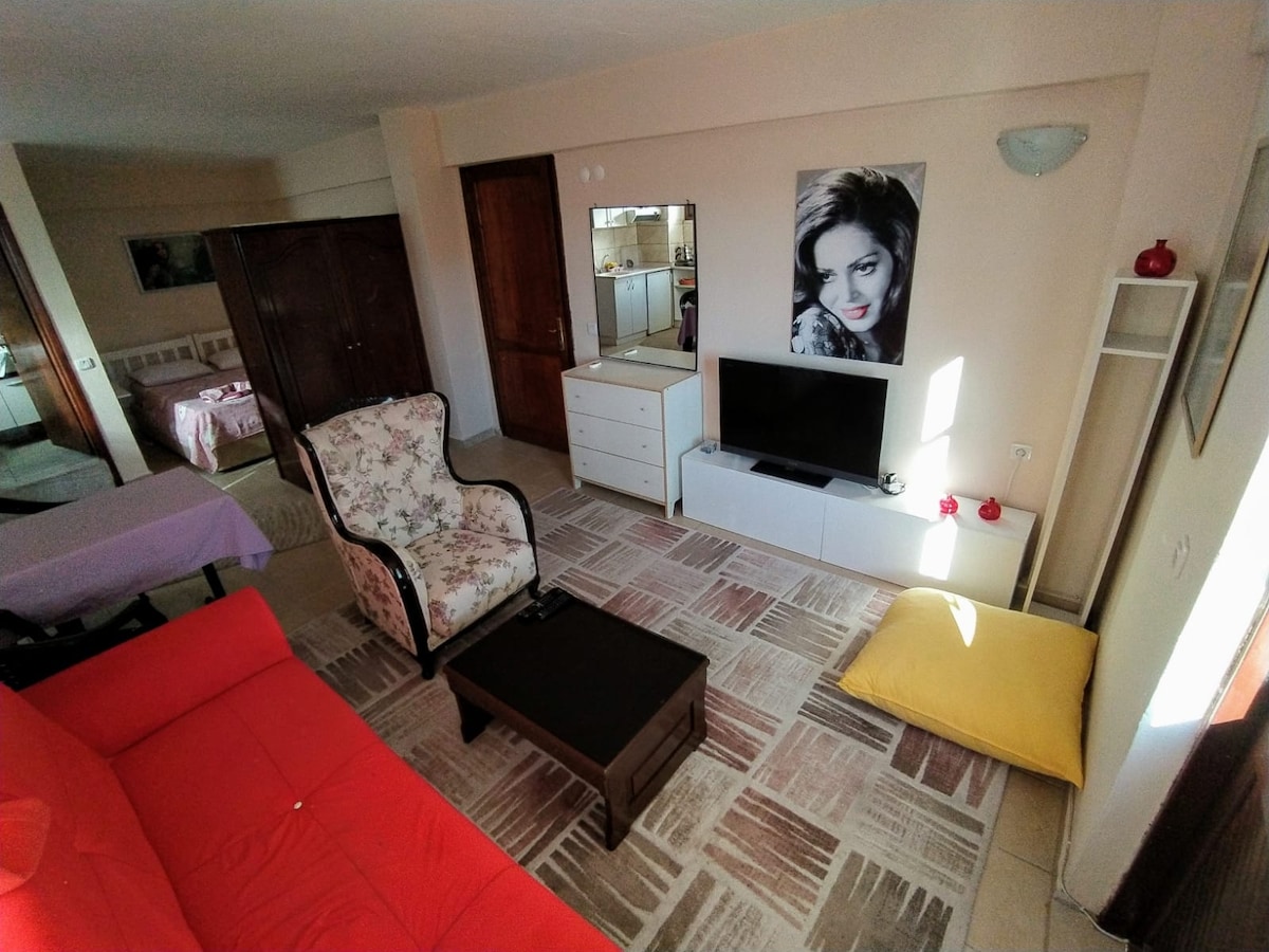 A spacious living area is furnished with a red sofa, a coffee table, and a decorative armchair. A TV is positioned on a white console, with a mirror reflecting the room. Natural light fills the space, complemented by soft textures in the decor.