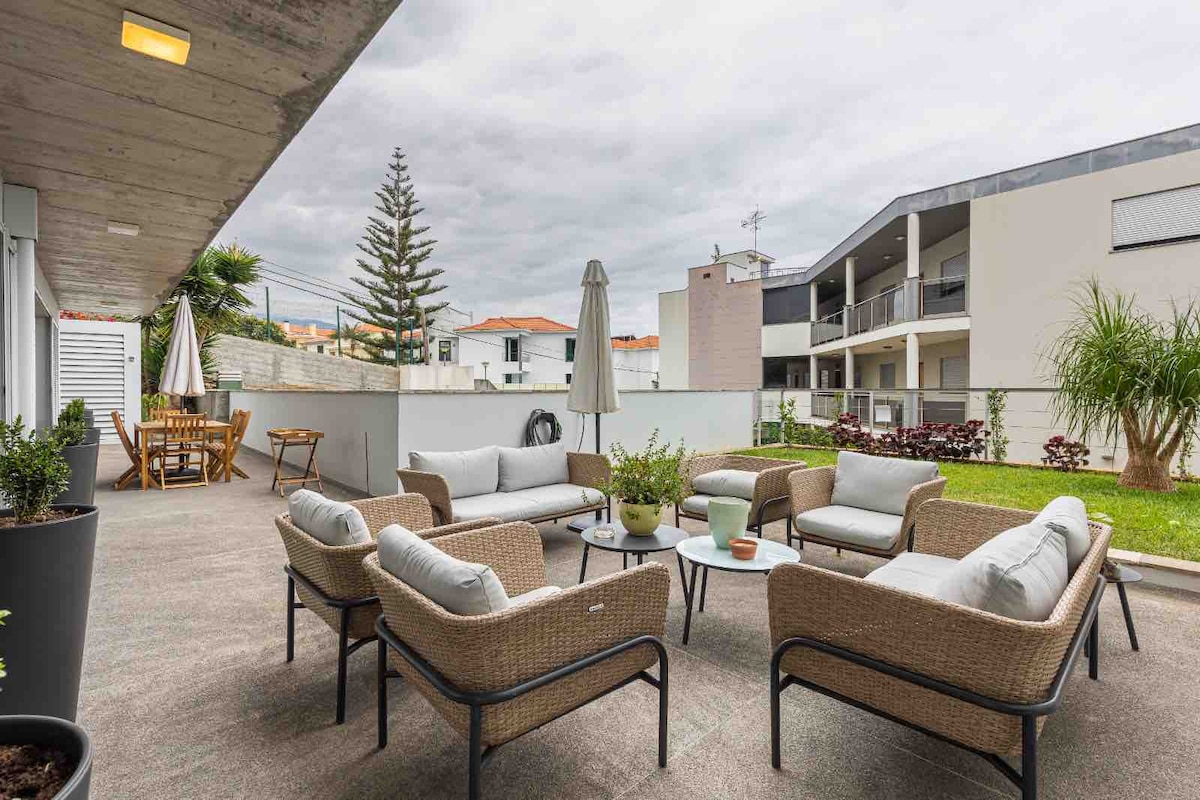 An outdoor patio area is visible, featuring a seating arrangement of light-colored cushioned chairs surrounding a small coffee table. A dining table is located in the background, with an umbrella providing shade. Signs of greenery appear alongside a small patch of grass.