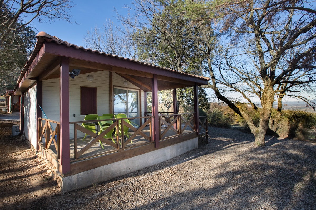 A chalet is positioned amid trees, featuring a covered terrace with green chairs and a table. The structure showcases large windows, allowing natural light to fill the interior. The surrounding ground is gravel, complemented by the natural landscape in the background.
