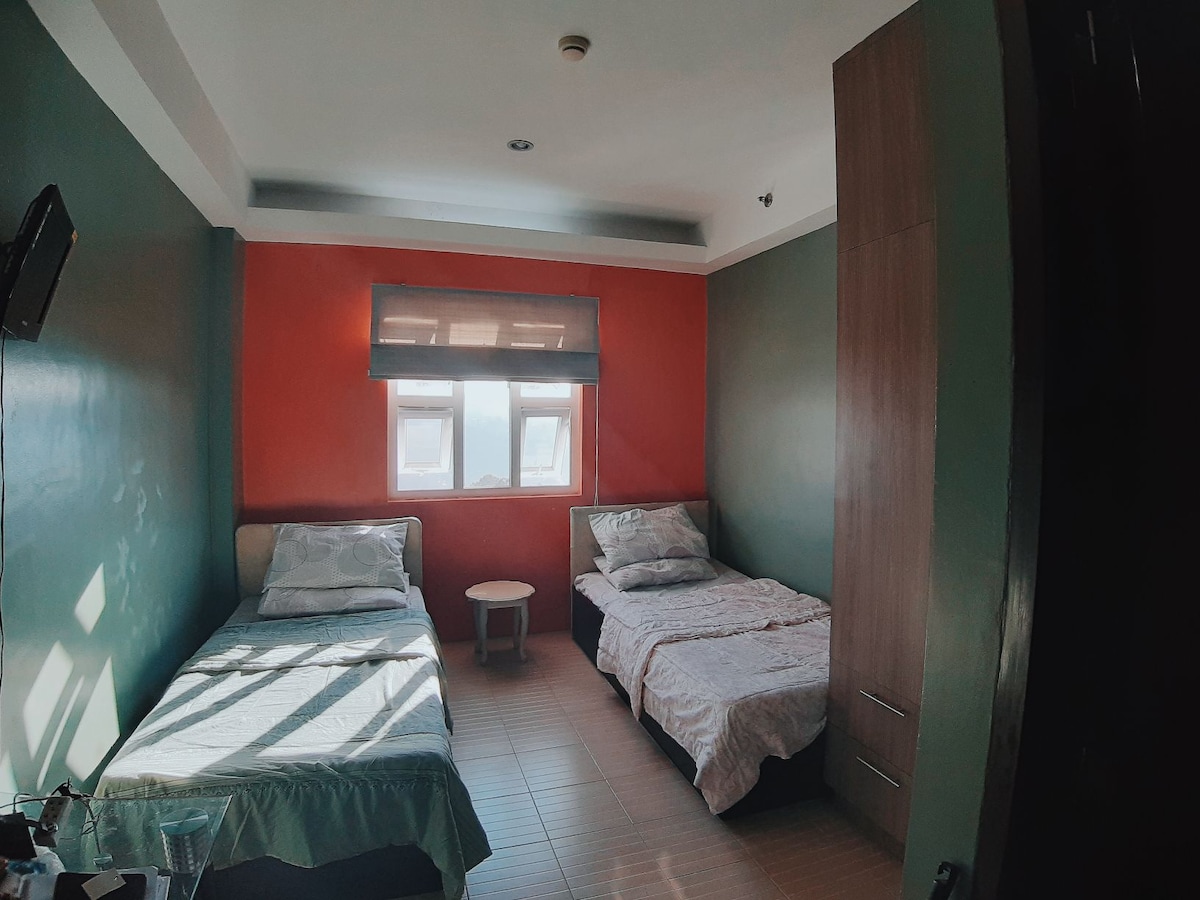 The room contains two beds with light bedding and decorative headboards. A small round table is positioned between the beds. The walls are painted in warm colors, providing a cozy ambiance. Natural light enters through two windows covered with shades.