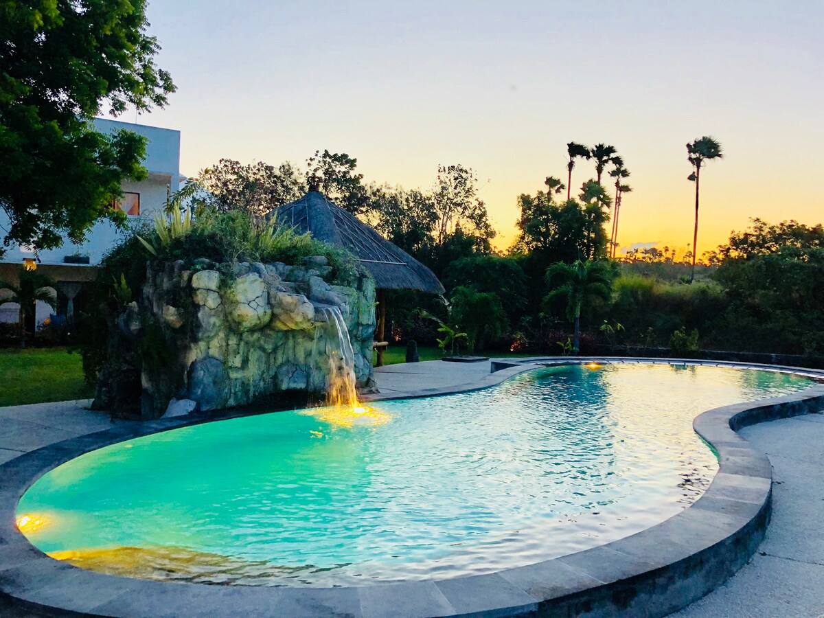 An inviting swimming pool is shaped organically, surrounded by lush greenery. A waterfall cascades from a rock formation into the pool, which is illuminated in soft turquoise hues as the sun sets in the background. A thatched roof structure is visible nearby.