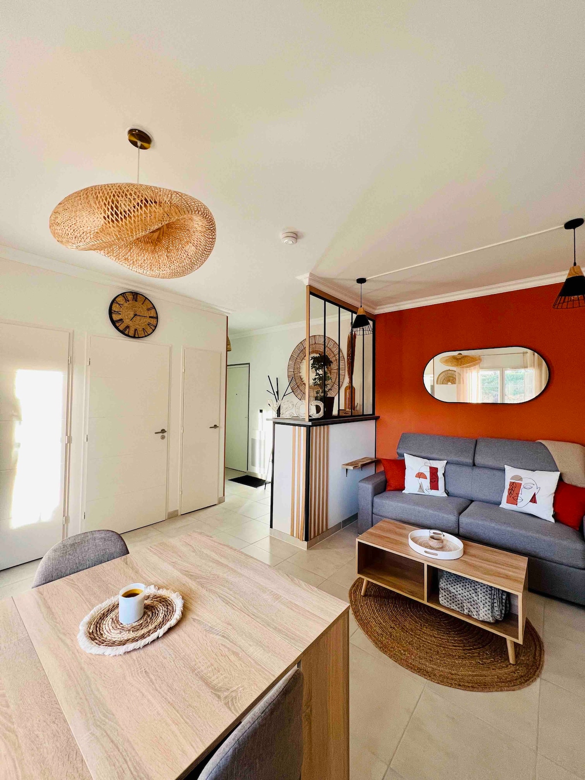 A cozy studio entrance showcases a stylish gray sofa against an orange accent wall. A wooden table is positioned nearby, accompanied by cushioned chairs. A decorative clock and mirror enhance the welcoming ambiance. Natural light filters through the doors, illuminating the space.
