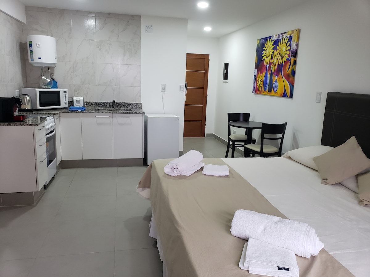 The image displays a comfortable studio apartment featuring a spacious bed with neatly arranged towels. A kitchenette is present, equipped with essential appliances and a dining area with two chairs. A colorful wall art piece adds a touch of vibrancy to the room.