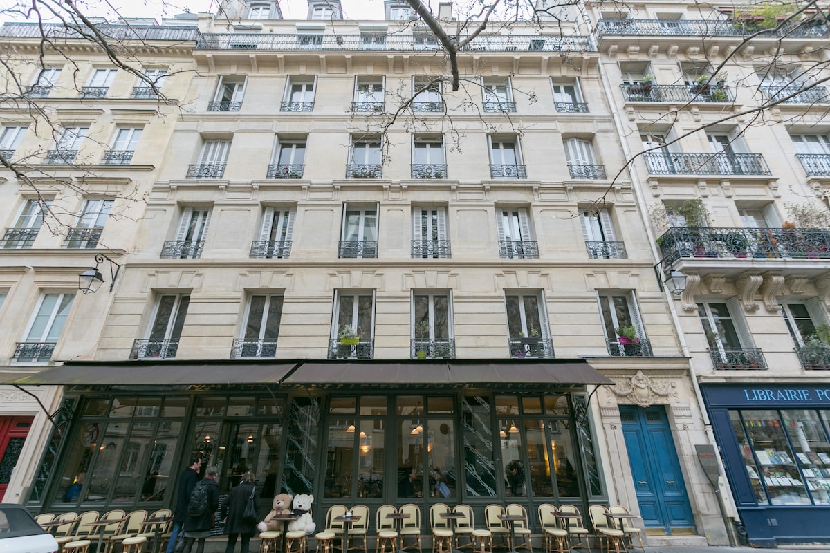 The apartment building features an elegant façade with multiple balconies adorned with greenery. Large windows provide ample natural light, while a charming café can be seen at street level, inviting guests to enjoy the lively Parisian atmosphere.