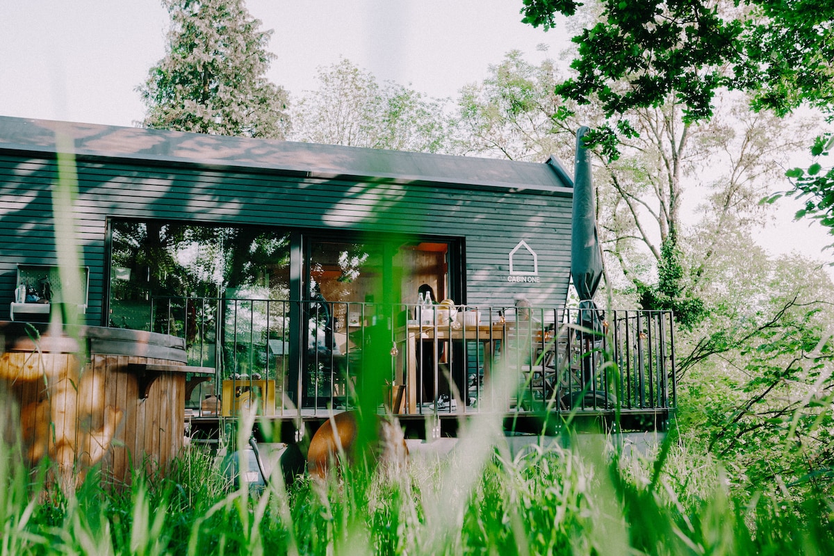 The cabin is showcased from a low angle, surrounded by lush green grass. Large glass doors open to a spacious deck, furnished for outdoor relaxation. A parasol stands beside the deck, providing shade, with trees framing the scene to create a serene atmosphere.