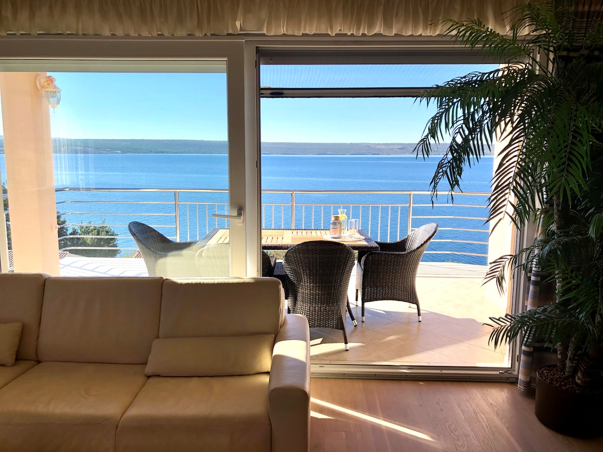A spacious living area is visible, featuring a soft sofa. Large sliding glass doors provide a view of a balcony equipped with outdoor seating. The calm sea stretches beyond, reflecting blue skies, creating a serene setting.
