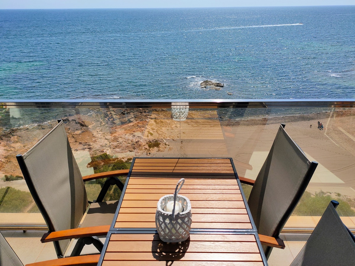 A balcony overlooking the sea features a wooden table surrounded by four metallic chairs. A decorative candle holder sits at the center. The tranquil waters are visible in the background, with rocky formations and a sandy beach below.