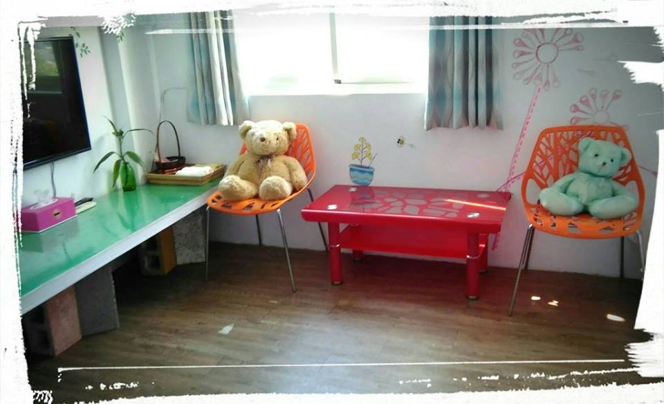A living area features a green tabletop, paired with two bright orange chairs adorned with plush toys. Natural light filters through sheer curtains, illuminating the space. A small potted plant and decorative wall art add a playful touch to the room.