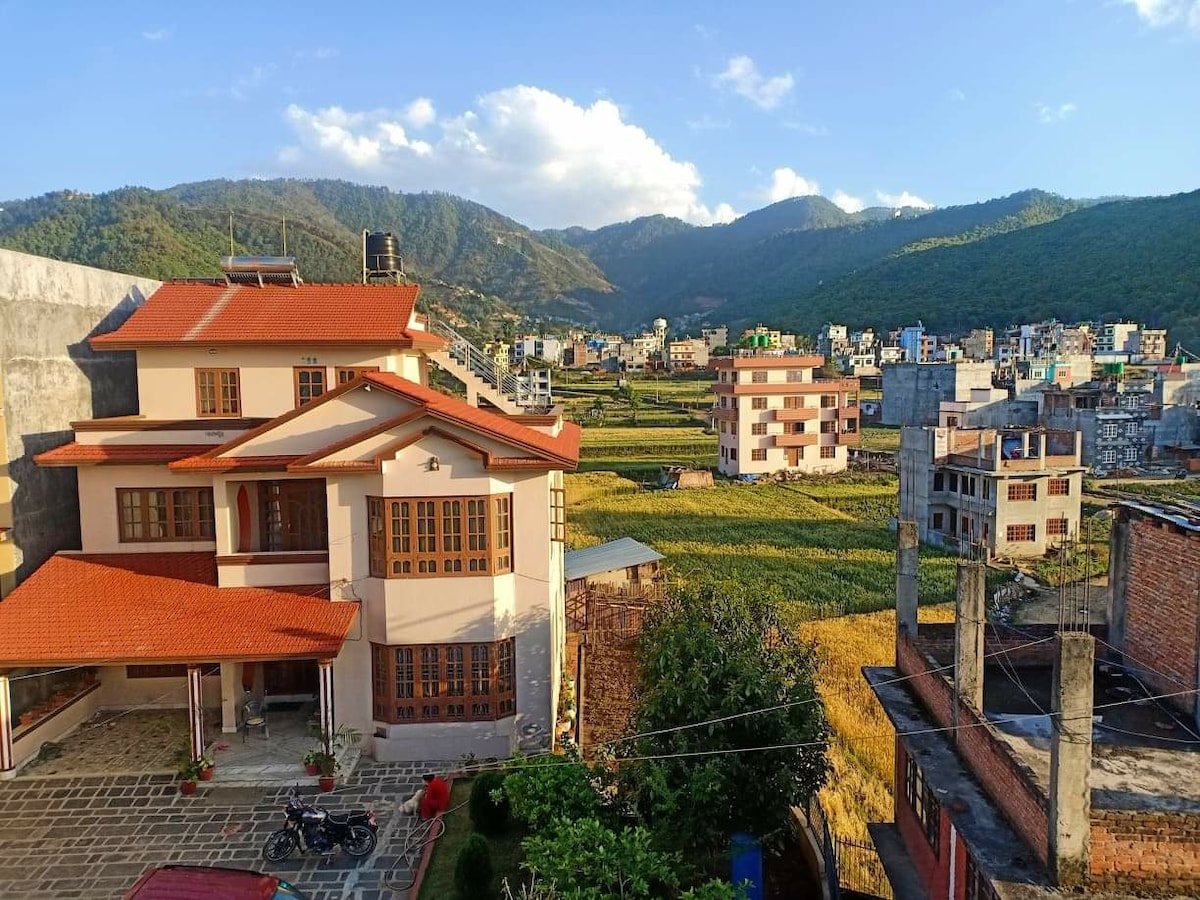 A multi-story building is set against a backdrop of rolling hills and vibrant fields. The red-tiled roof contrasts with the expansive green landscape, while nearby structures create a sense of community. The image captures the serene environment of the Kathmandu valley.