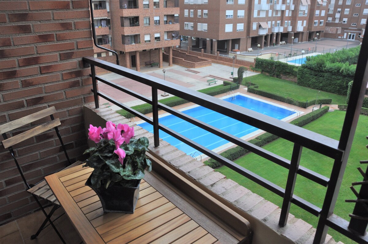 A balcony view features a small wooden table and metal chair, adorned with a potted pink flower. The surrounding area includes manicured lawns and two community swimming pools, complementing the modern brick buildings in the background.