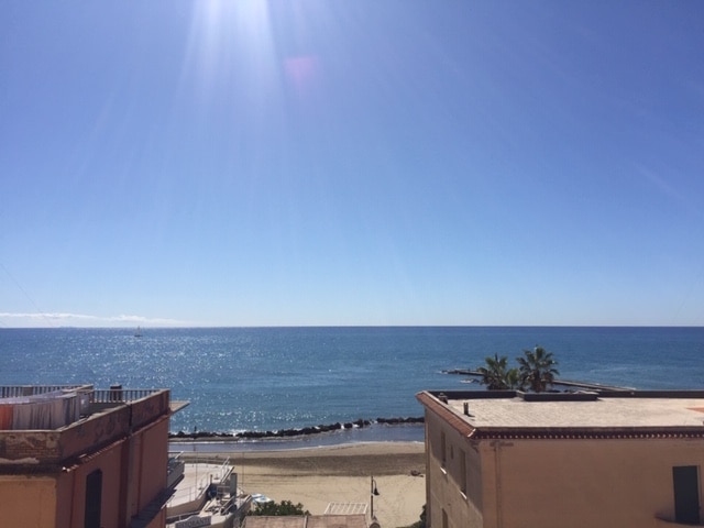 A clear view of the tranquil sea extends to the horizon, reflecting sunlight in shimmering patterns. The sandy beach is visible along the shoreline, with coastline structures framing the scene. Palm trees are observed in the foreground, contributing to the beachside ambiance.