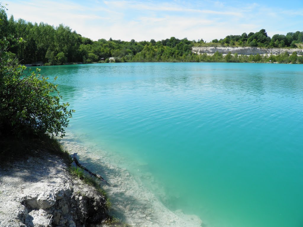 A serene lake features clear turquoise water surrounded by lush greenery. The rocky shoreline gradually slopes into the water, highlighting the contrast between the vibrant blues and the natural earth tones. Tree-lined banks enhance the peaceful atmosphere of this outdoor space.
