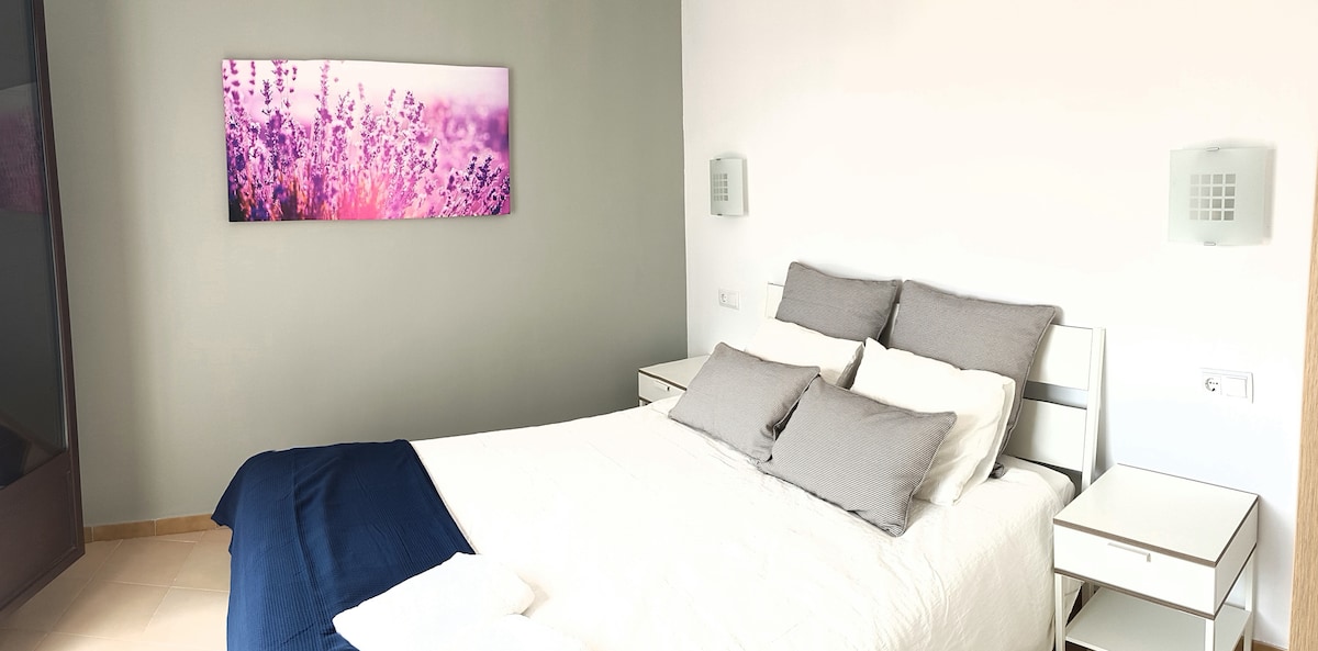 A serene bedroom features a spacious bed adorned with soft, neutral-toned pillows. Two bedside tables hold minimalist lamps, and a wall is decorated with a vibrant nature-themed artwork. Natural light enhances the bright, calming color scheme of the room.
