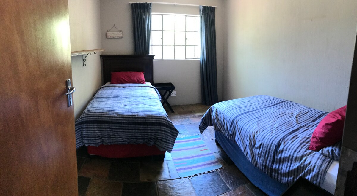 A room is presented with two beds, each covered in striped blue bed linens and accented with red pillows. A window allows natural light to illuminate the space, and a small, dark wood table is positioned beside the beds. The flooring features a combination of tiles in earthy tones.