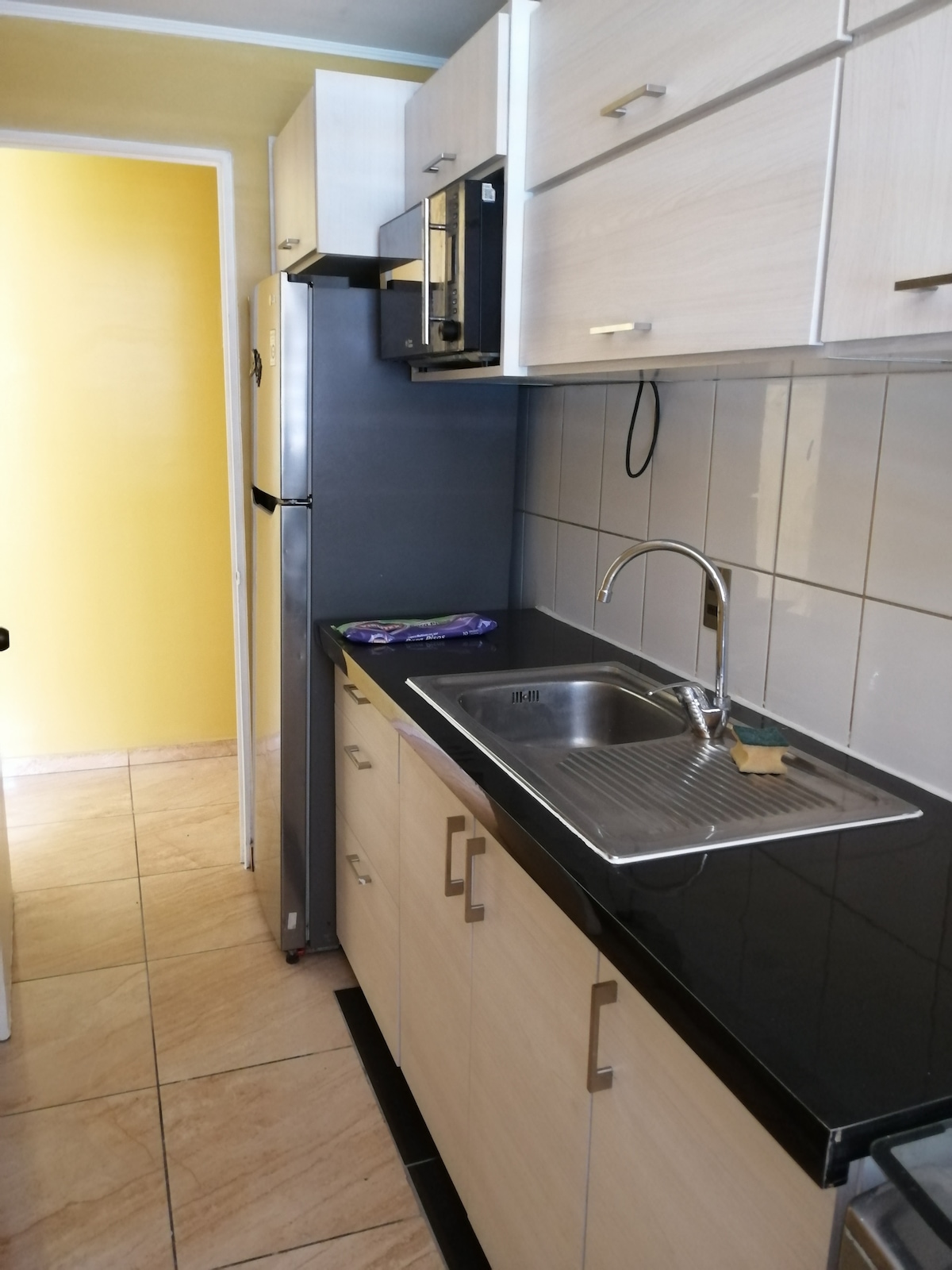 A compact kitchen area features modern cabinetry with a light wood finish and dark countertops. Stainless steel appliances, including a refrigerator and an oven, are visible. A single basin sink is integrated into the counter, with light tile backsplash providing a clean look.