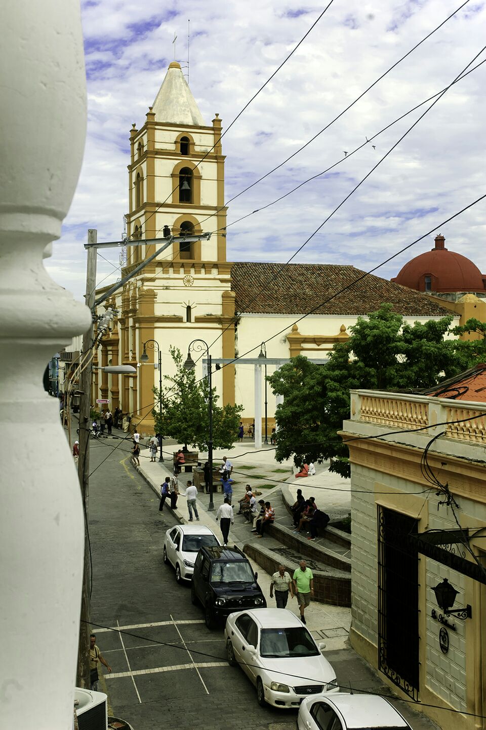 A view from a balcony overlooks a bustling street featuring a historic church with a tall bell tower, surrounded by lush trees and vibrant activity. People are seen walking along the street, with cars parked nearby, conveying a lively urban atmosphere.