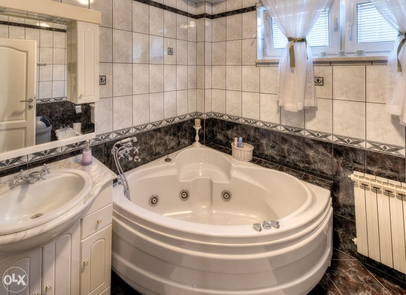 A spacious bathroom is equipped with a corner jacuzzi tub, surrounded by elegant tiled walls in various shades of gray. A sleek vanity with a sink is featured, alongside a large mirror. Natural light filters through windows adorned with light drapes.