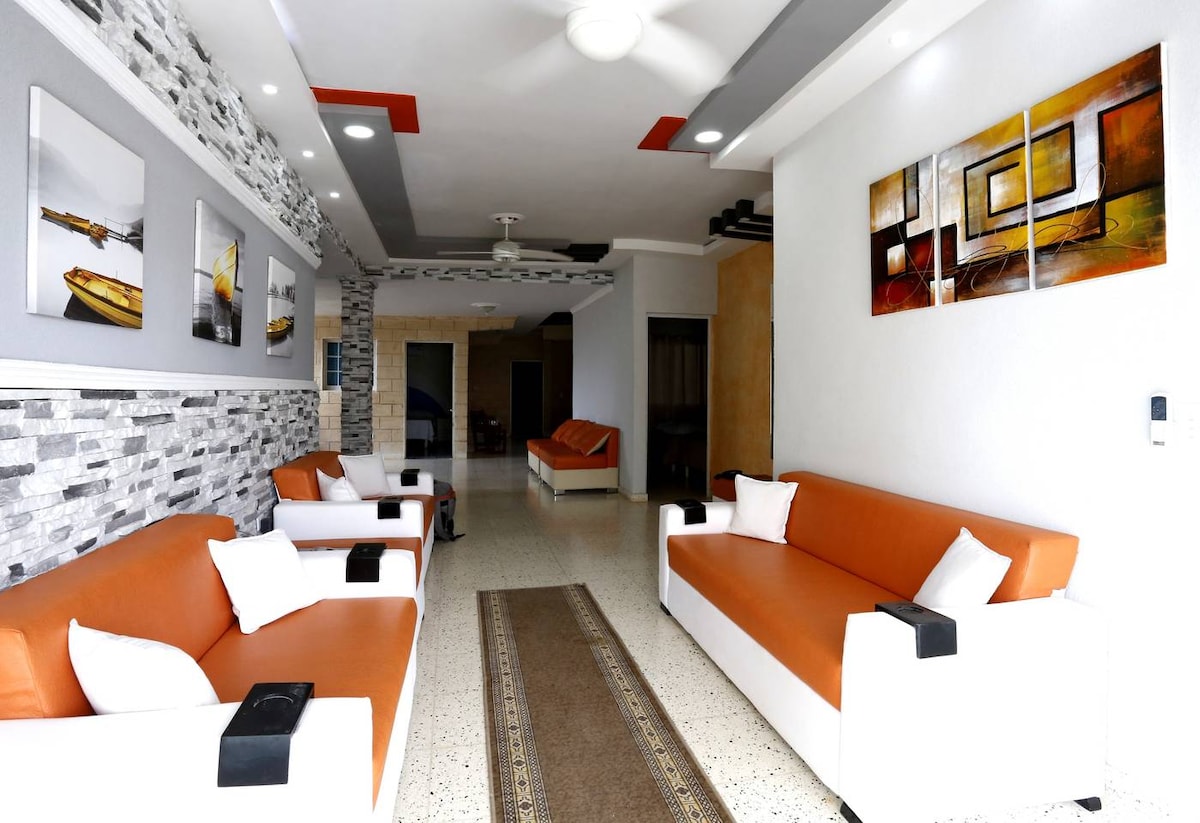 A modern common area features two orange sofas with white armrests, complemented by decorative pillows. The space showcases textured stone walls, a stylish area rug, and abstract artwork. Ceiling fans provide airflow, while recessed lighting enhances the overall bright and welcoming environment.