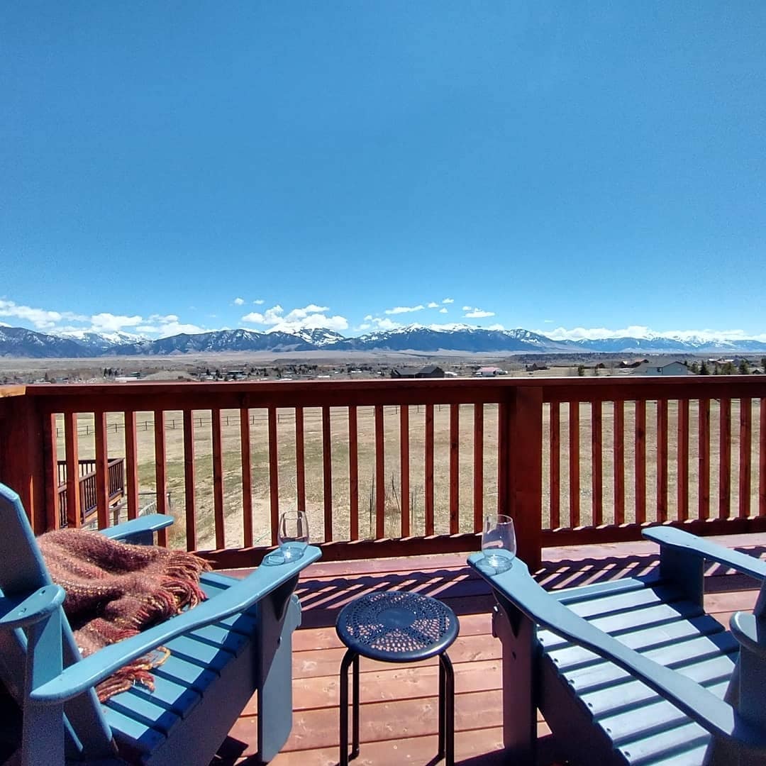 A spacious deck is depicted with two blue Adirondack chairs and a small round table, offering expansive views of the mountains under a clear blue sky. Light stretches across the deck's wood surface, enhancing the natural landscape.