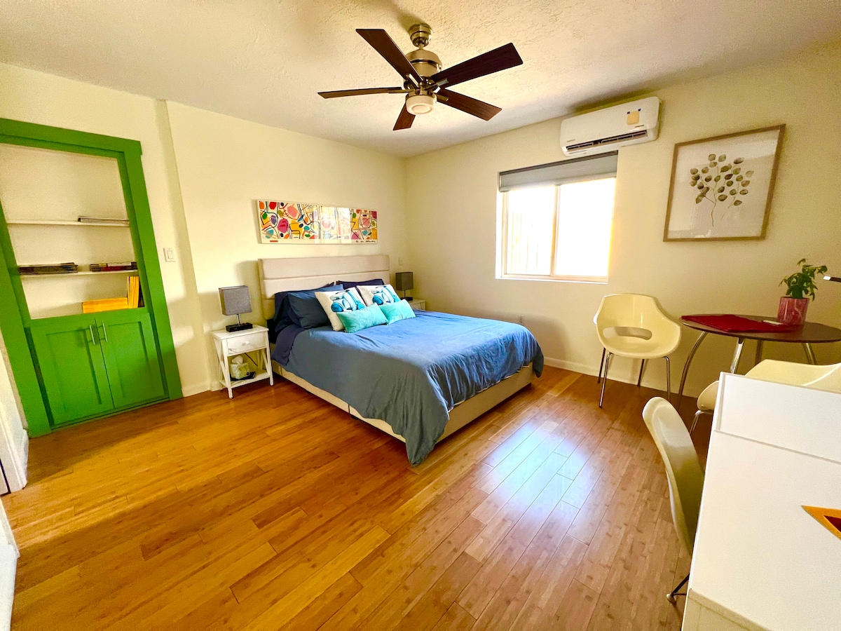 A spacious studio features a comfortable bed adorned with colorful pillows. A large window allows natural light to fill the room. The space includes a small desk, a chair, and a built-in shelf with a vibrant green accent cabinet.