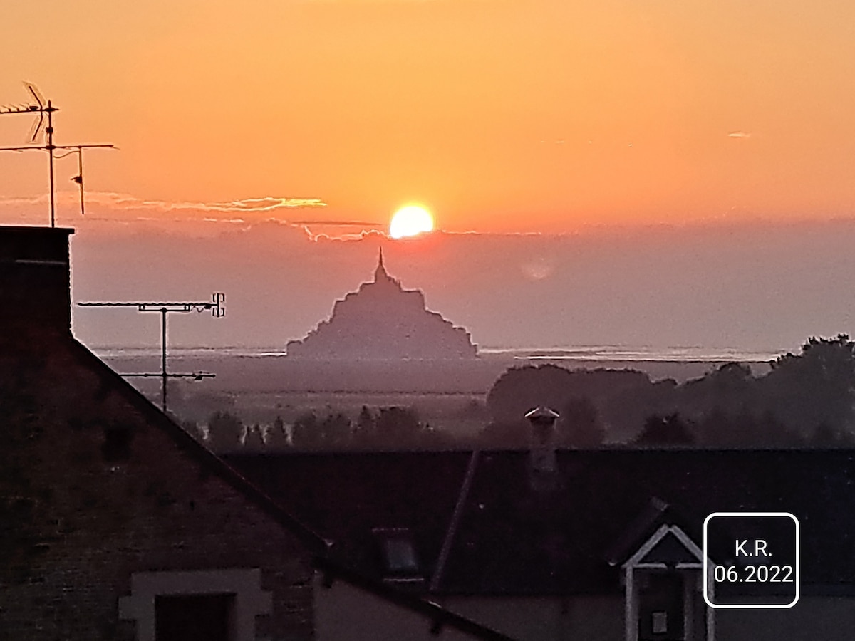 A sunset is captured over the silhouette of a historic structure, likely Mont Saint-Michel, with the sun positioned just above the horizon. Soft colors blend in the sky, creating a tranquil evening ambiance. Rooftops and antennas are visible in the foreground.