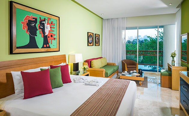 A vibrant guest room is highlighted by green walls and artistic wall decor. A spacious bed with accent pillows sits alongside a seating area. Natural light filters through large sliding doors, revealing a private terrace with views of greenery.