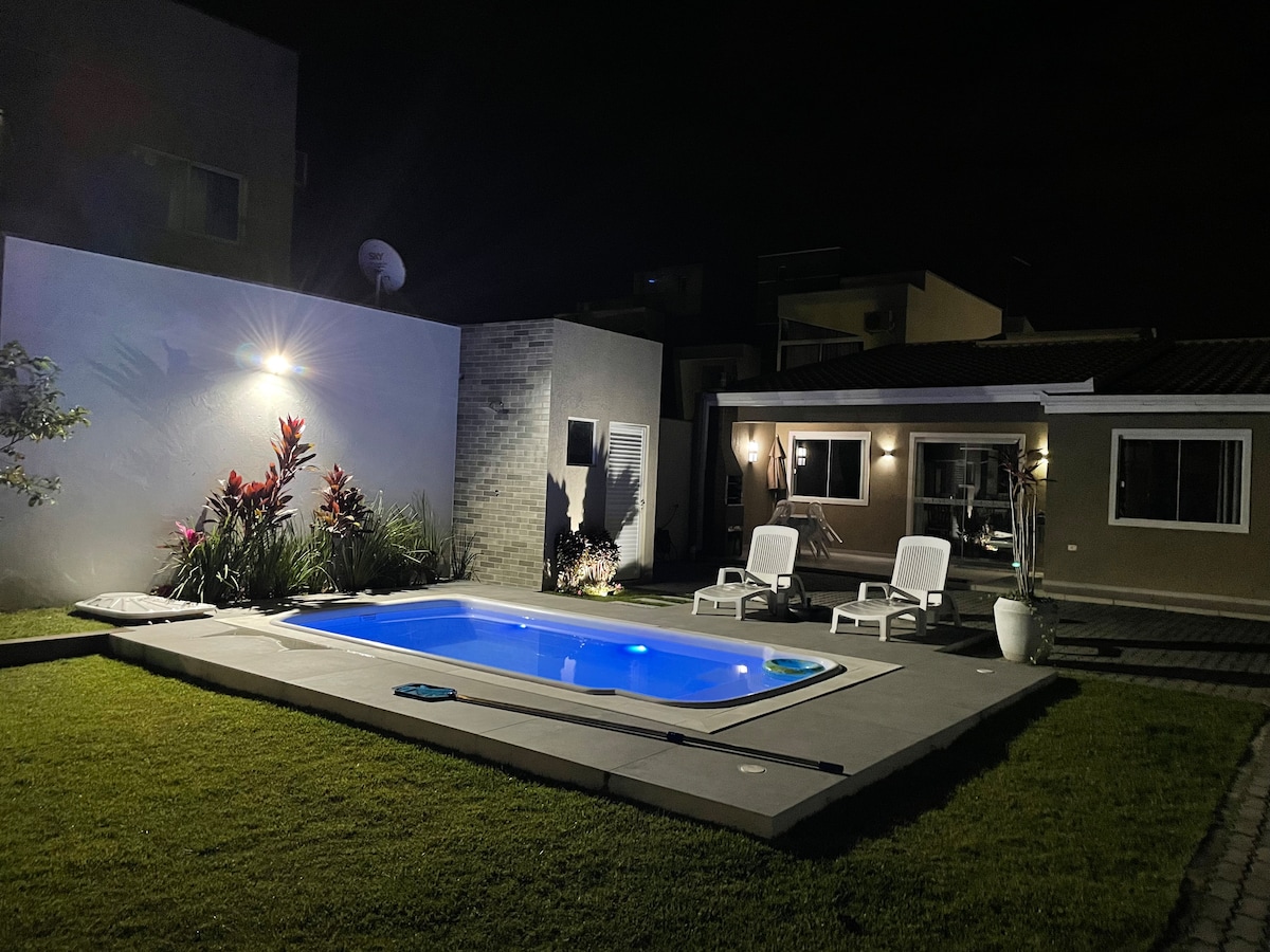 The tranquil pool is illuminated in blue light, surrounded by a neat concrete patio. Two lounge chairs are placed nearby, offering a space for relaxation. Lush plants enhance the ambiance, while the house features large windows reflecting the night sky.