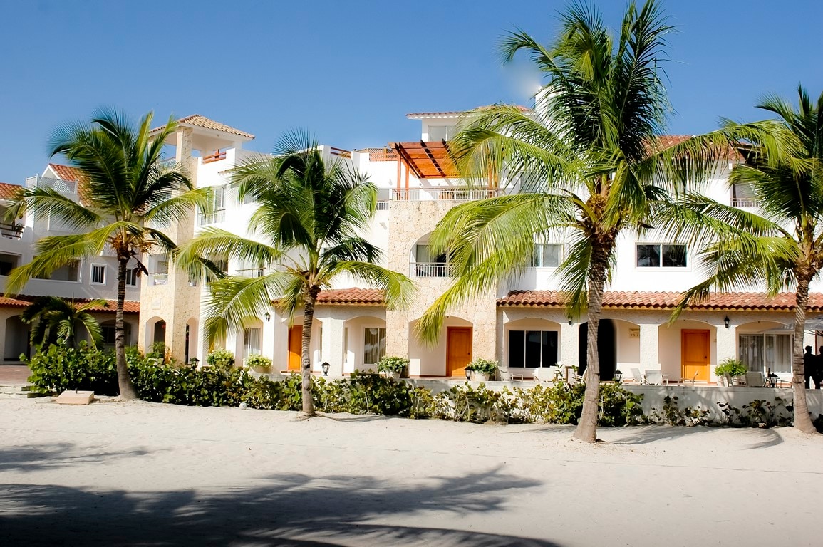A resort-style building is surrounded by swaying palm trees, with sandy pathways leading to the sandy beach. The white-stucco structure features multiple balconies and terraces, providing access to outdoor spaces. Lush greenery is visible along the base, enhancing the tropical ambiance.