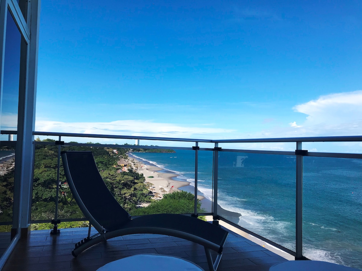 A shaded lounge chair is positioned on a private balcony, offering expansive views of the coastline and the ocean. The blue sky merges with the water, creating a serene backdrop. Lush greenery is visible along the shore, enhancing the natural beauty of the setting.