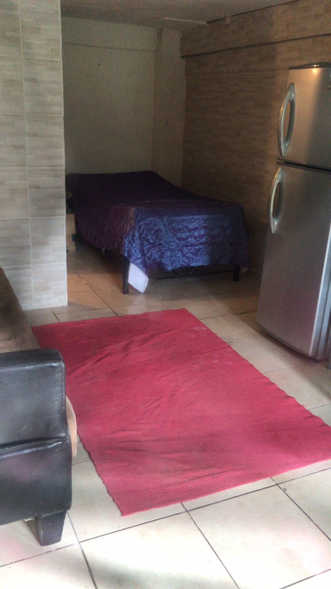 An interior space is visible, featuring a single bed covered with a purple blanket in the corner. A red rug lies on the tiled floor, contrasting with the neutral-colored walls. Nearby, a silver refrigerator stands next to a dark sofa, creating a compact living area.