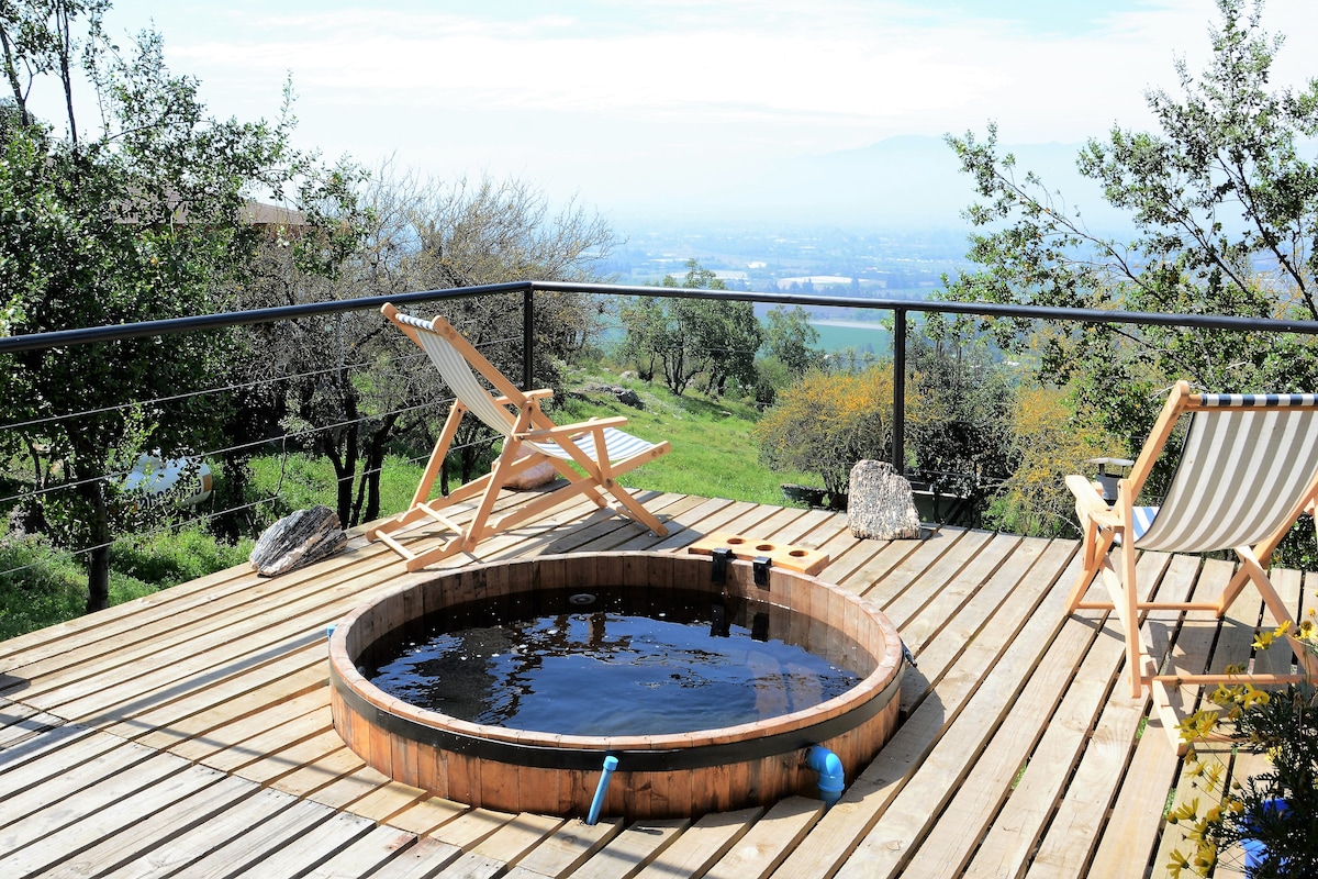 A wooden hot tub is positioned on a wooden deck, offering scenic views of the valley below. Two reclining chairs are placed nearby, surrounded by greenery. The landscape features rolling hills and distant mountains under a clear sky.