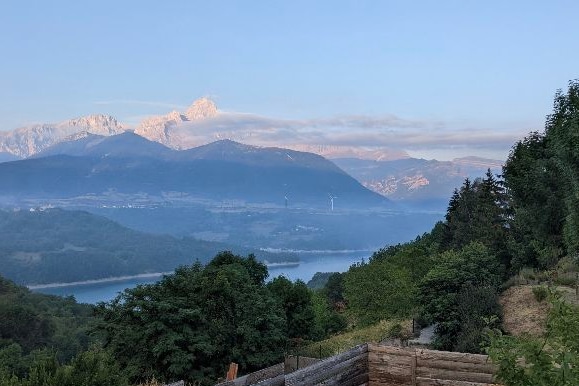A panoramic view captures the serene landscape, featuring a lake bordered by lush greenery. In the distance, majestic mountains rise under a soft blue sky, illuminated by morning light, creating a tranquil setting.