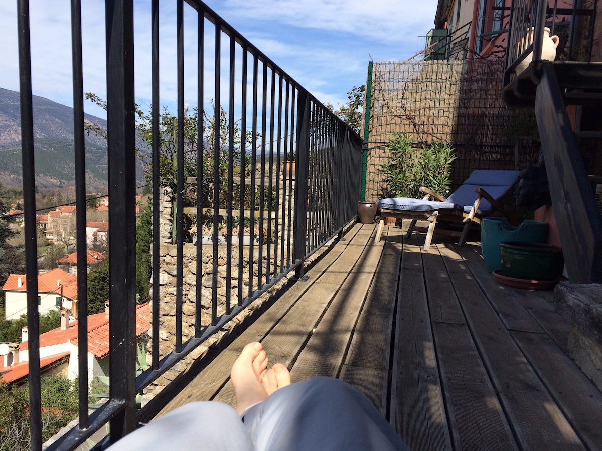 A spacious wooden terrace is visible, overlooking a scenic valley with distant mountains. Sunlight casts shadows across the deck, highlighting lounging chairs and potted plants. The peaceful atmosphere invites relaxation, with neighboring rooftops and hills framed in the background.