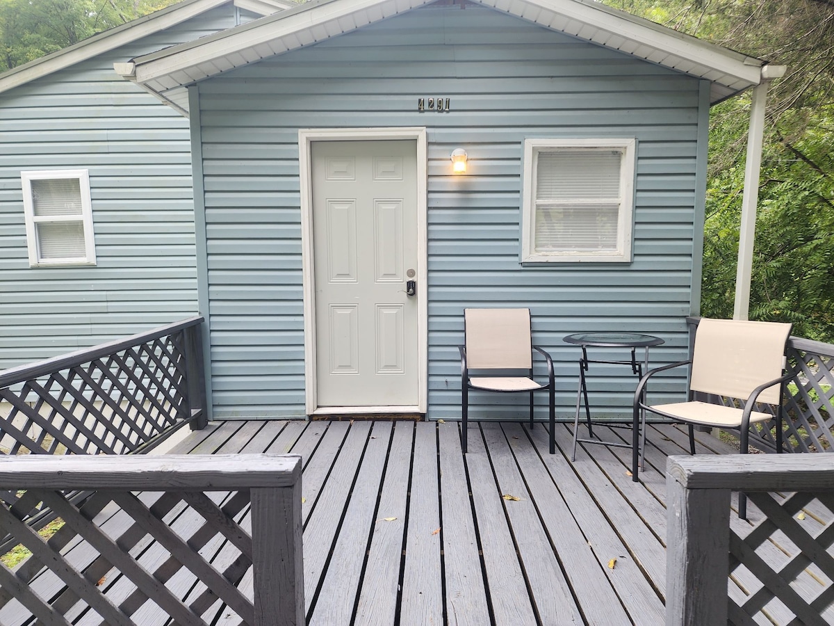 A blue exterior cottage is featured with a white door, flanked by two light-colored chairs set around a small table on a wooden deck. Latticework surrounds the deck, adding privacy and charm. Natural greenery is visible in the background.