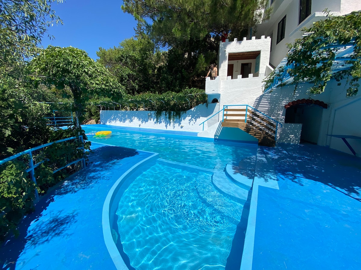 An inviting swimming pool, surrounded by lush greenery, features vibrant blue water reflecting the clear sky. Steps lead to a recessed area, while a path lined with blue railings connects the pool to the cottage and outdoor seating areas.